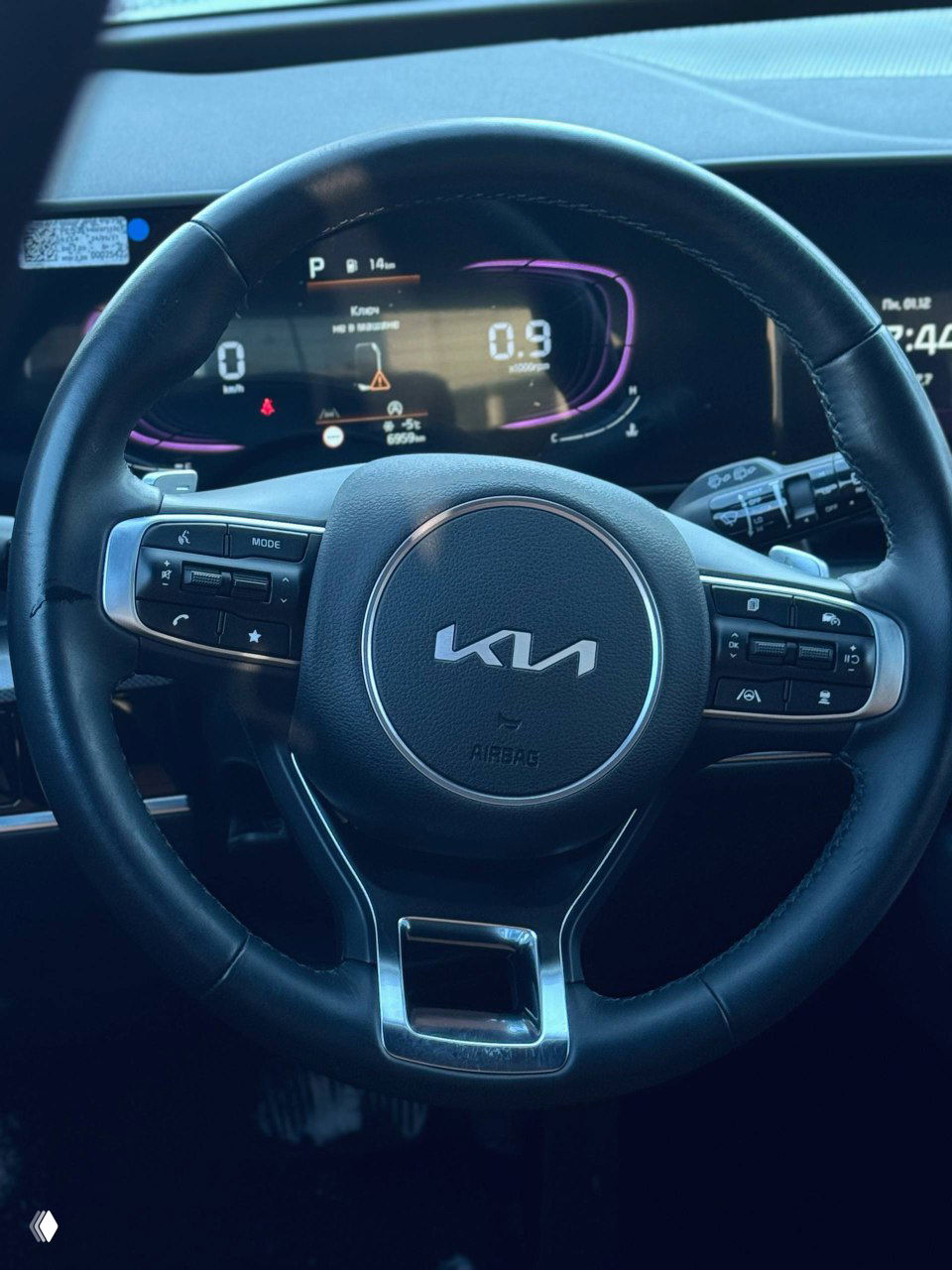Руль KIA с крупным логотипом на подушке безопасности, на фоне цифровой приборной панели и мультимедийного экрана видны элементы управления