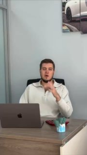 Мужчина за столом с ноутбуком в офисе TopCar объясняет вопросы растаможки авто; на стене видна часть фото автомобиля.