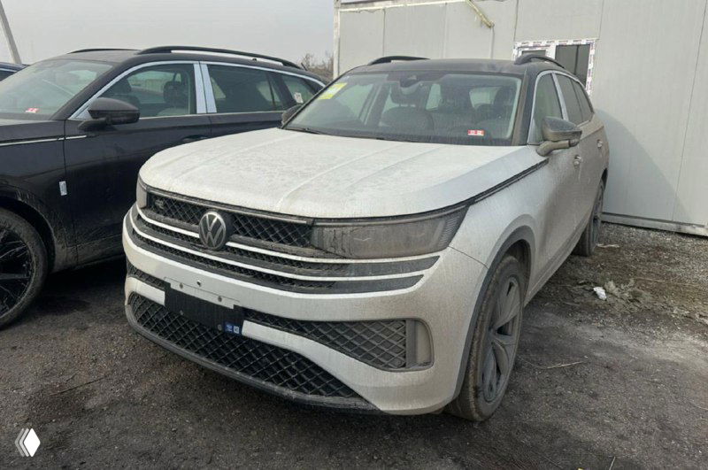 Грязный белый Volkswagen Tavendor на стоянке — автомобиль после перевозки, покрыт дорожной пылью, вид спереди-слева, рядом другие машины