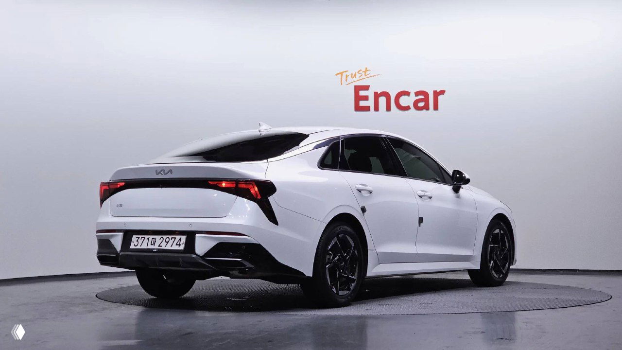 KIA K5 2024, вид сзади в студии: белый кузов, видны задние фонари и бампер, автомобиль на поворотном подиуме для показа состояния.
