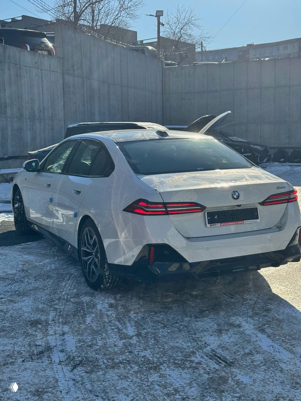 Задний ракурс белого BMW 520i с видимыми фонарями, багажником и линиями кузова, автомобиль стоит на зимнем асфальте у бетонной стены.