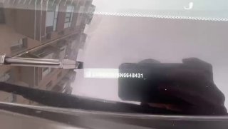 Фотография фрагмента лобового стекла с видимым идентификационным номером кузова (VIN), снята крупным планом на парковке у здания.