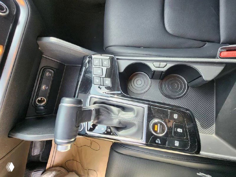 Слева крупный план селектора АКПП и центральной консоли KIA Sportage 2024 с элементами управления и подстаканниками, отделка в чёрном цвете.