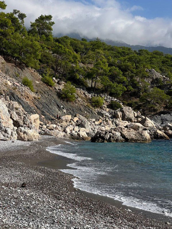 Побережье с кромкой прибоя на гальке, спокойная морская гладь и лесистые склоны вдалеке — детали пляжного берега Ликийки весной или осенью.