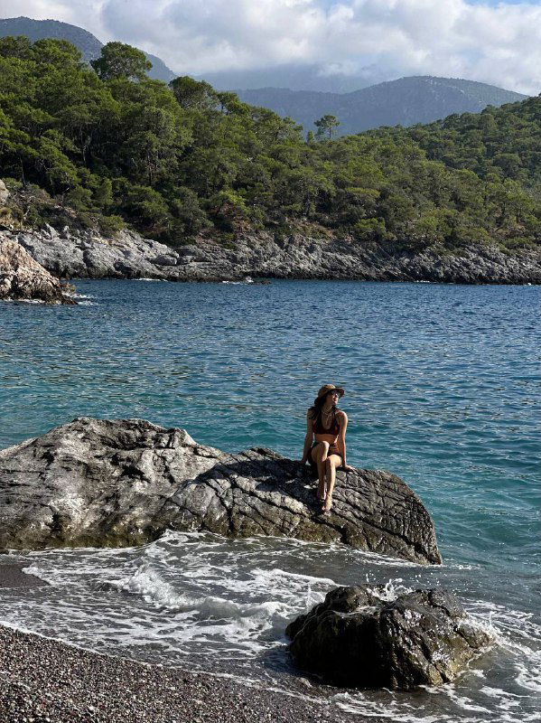 Человек сидит на крупном камне у моря, вид на спокойную бухту и горы на фоне — момент отдыха после похода на берегу Ликийки.