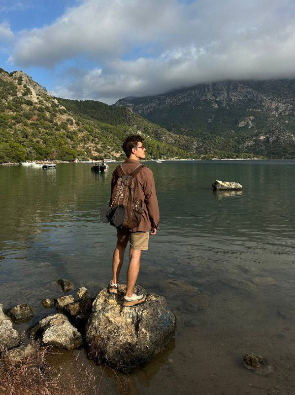 Пейзаж с человеком на камне и тучами над горным хребтом, сочетание спокойной воды и драматичного неба — вечерние мотивы маршрута.