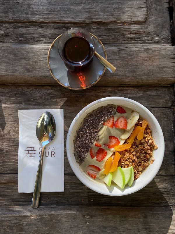 Смузи-боул с фруктами и гранолой на деревянном столе рядом с чашкой чая и салфеткой с логотипом Casa Sur — завтрак в саду.