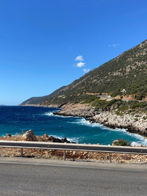 Береговая линия с металлическим ограждением на краю трассы, бирюзовое море и скалы под голубым небом — отрезок пути вдоль побережья.