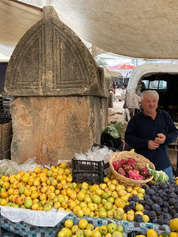 Прилавок турецкого рынка с множеством жёлтых фруктов, виноградом и корзиной сладостей; продавец сидит под тентом рядом с товаром и улыбается