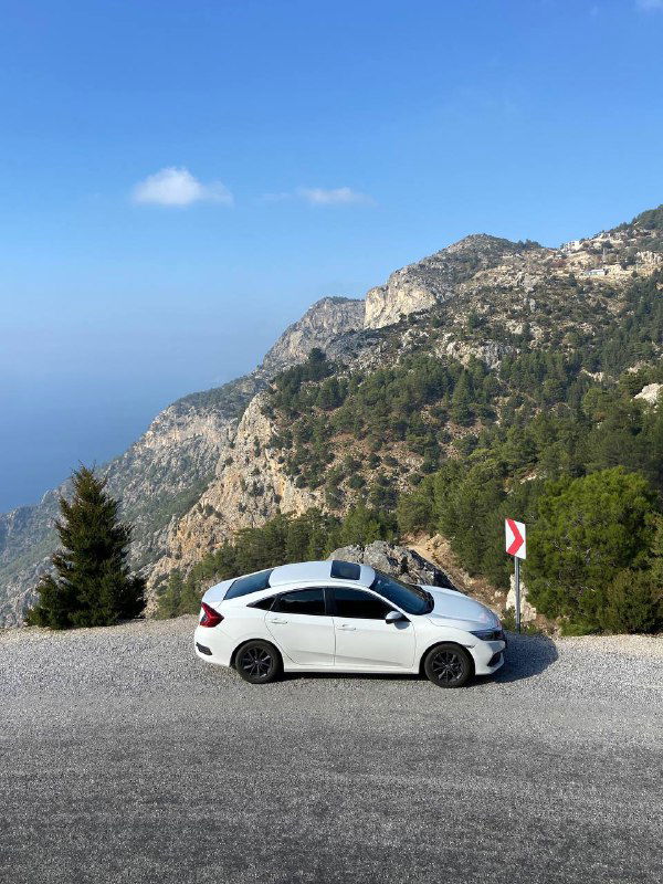 Белый автомобиль припаркован на узкой горной дороге без ограждений, вокруг скалы и обрыв, видна часть побережья и ясное небо.