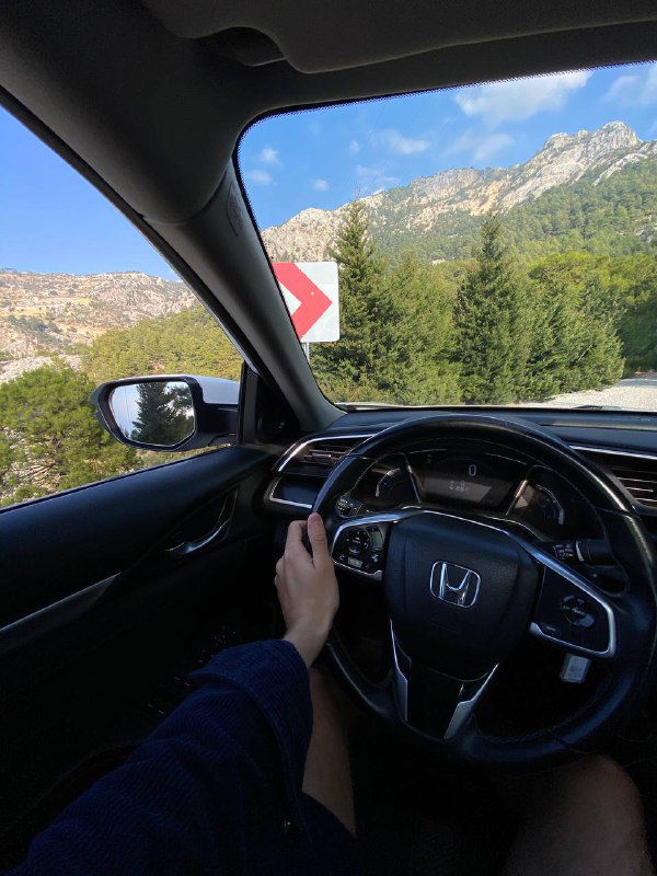 Руки водителя на руле внутри автомобиля, через лобовое стекло видна извилистая горная дорога, скалы и зеленые склоны — поездка по серпантину.