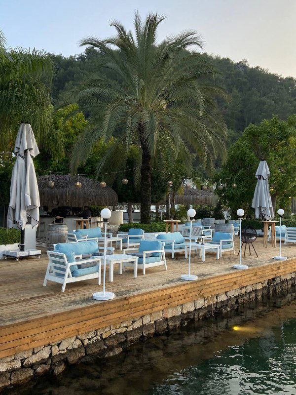 Набережная и зона отдыха Mori Restoran: голубые диваны и белая мебель у воды, пальмы и зелёные холмы на фоне курорта Фетхие.