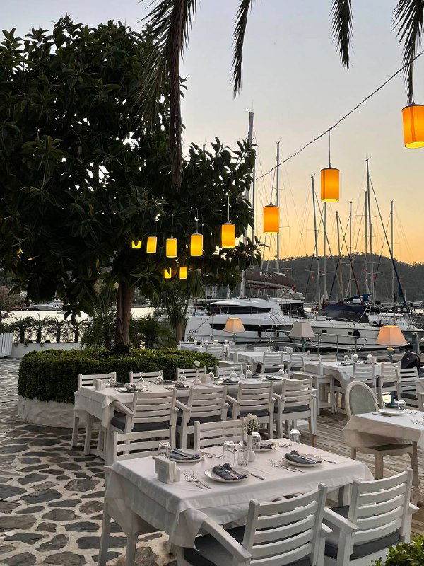 Вечерняя терраса Mori Restoran: ряд белых столов вдоль мачт яхт, тёплое освещение подвесных ламп и пейзаж марина на закате.