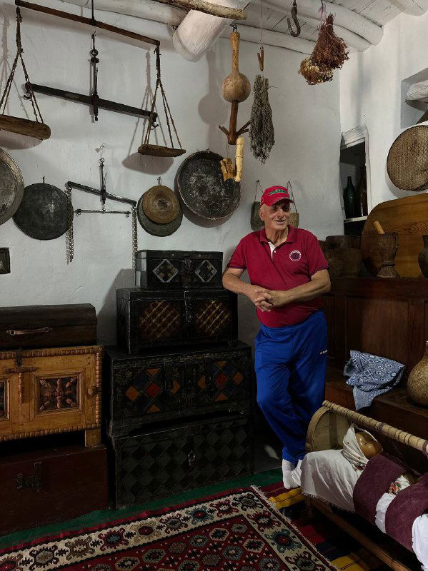 Магомед в комнате с коллекцией старинных предметов: музыкальные инструменты, сундуки и деревянный интерьер этно‑сакли.