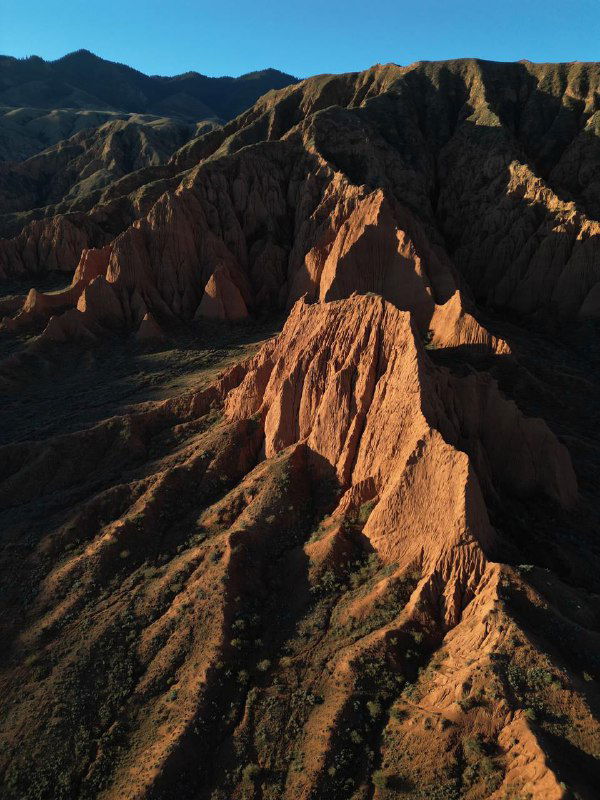 Красные каньоны Кыргызстана с глубокими складками и острыми хребтами на фоне синего неба, воздушная фотография подчёркивает марсианскую текстуру ландшафта.