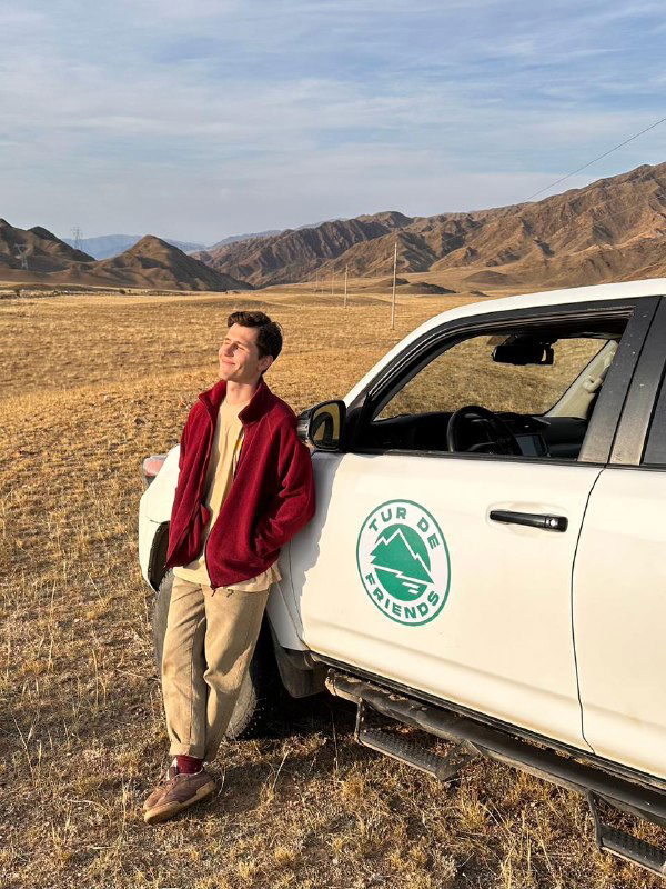 Путешественник у внедорожника с логотипом Tur de Friends на фоне открытой равнины и гор — часть экспедиции и логистика поездки.