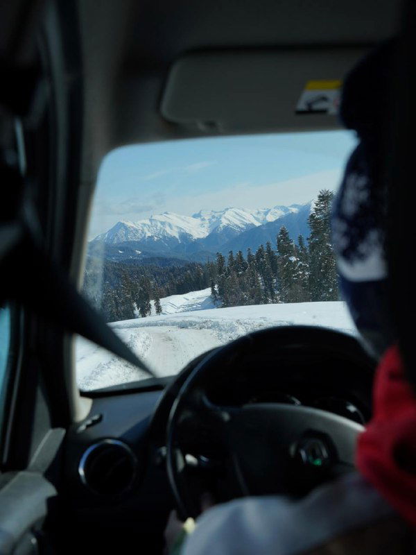 Вид из салона автомобиля на заснеженную дорогу и отдалённые горные пики, пассажир на переднем плане, путь к курортным зонам.