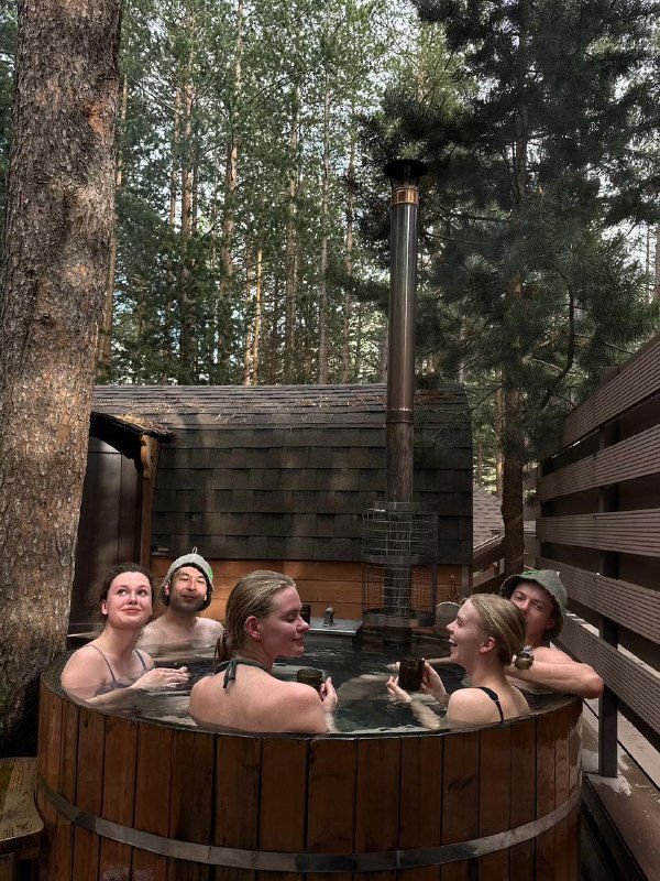Компания отдыхающих в деревянной фурако‑ванне на открытом воздухе среди соснового леса, пар над водой и тёплая дружеская атмосфера.