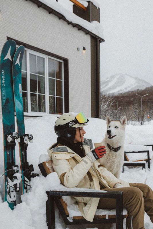 Отдыхающий в зимней одежде с хаски на шезлонге у домика, снег вокруг и уютная база отдыха на фоне склонов и деревьев.