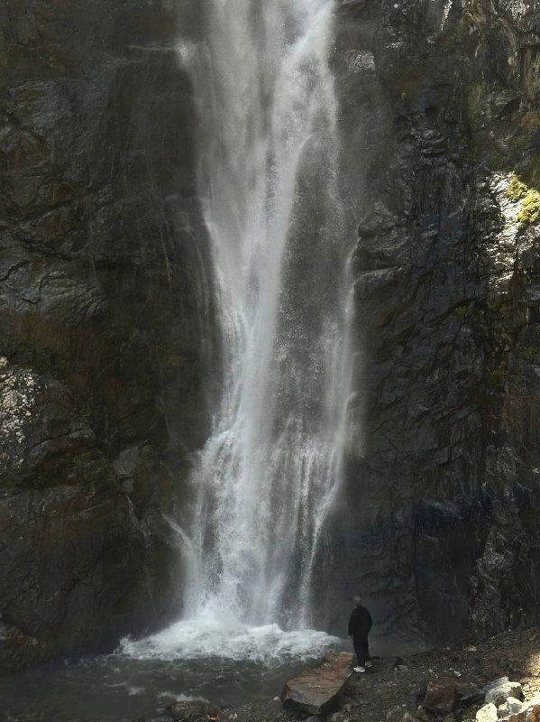 Высокий водопад в ущелье: вода стремительно стекает по скалам, у подножия виден человек для масштаба, окружение каменистое и зелёное.