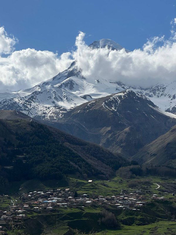 Заснеженная гора над деревней Степанцминда: панорама с домами в долине и крутыми склонами под голубым небом.