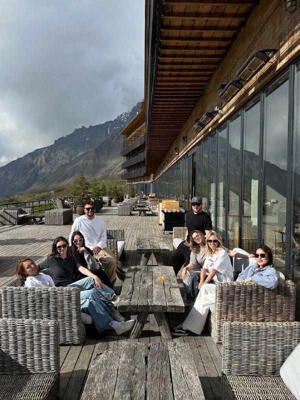 Гости на просторной террасе отеля: группа людей отдыхает на плетёной мебели с видом на горы и долину Степанцминды.
