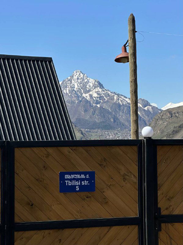 Деревянный забор с адресной табличкой «Tbilisi str.» на переднем плане и высокие вершины Кавказа на заднем плане под облачным небом.