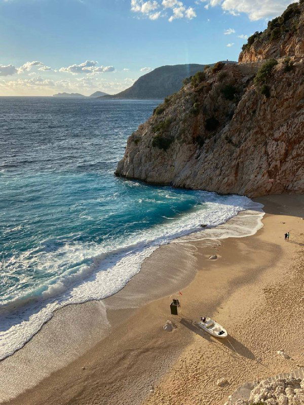 Пустынный галечный пляж у крутого скального берега, бирюзовая вода и небольшая лодка на песке — типичный вид средиземноморского побережья.