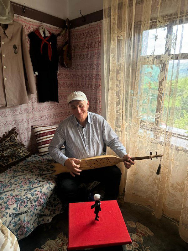 Местный музыкант в доме в ауле: мужчина с традиционным инструментом на фоне текстиля и уютной комнаты, живой быт гостей.