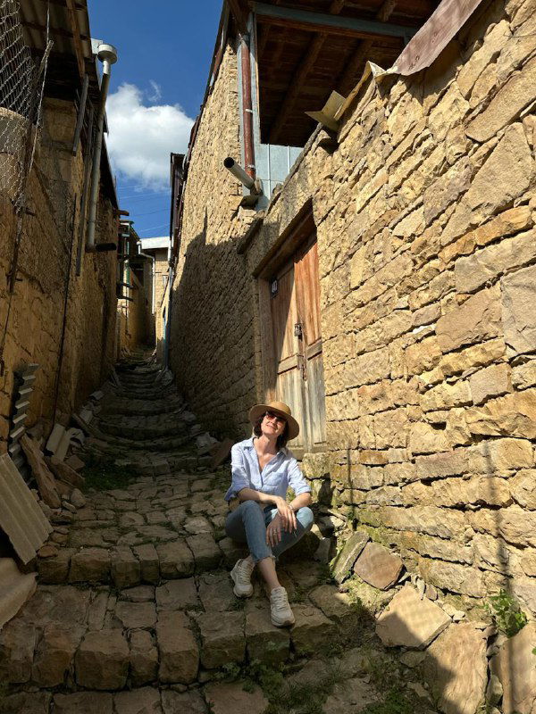 Узкая каменная улочка в ауле с высокими стенами и сидящей путешественницей — атмосферная сцена деревенской жизни и пейзажа.