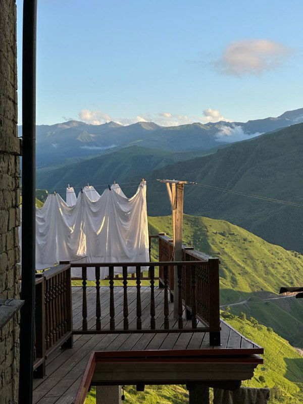 Балкон кафе с развешенным белым бельём, деревянными перилами, видом на красные крыши аула и зелёные горные террасы на горизонте.