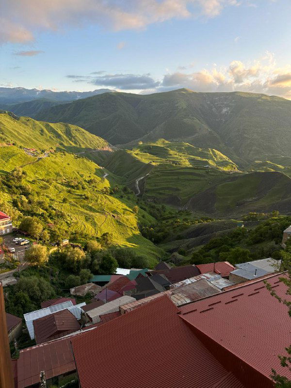 Панорамный вид с высоты: красные крыши аула на переднем плане, вдалеке извилистая дорога и зелёные горные поля.