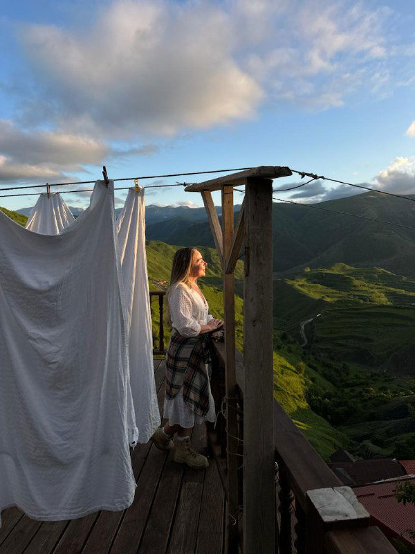 Женщина на деревянной террасе кафе рядом с развешенным бельём, смотрит на зелёные горные склоны и долину при закате.