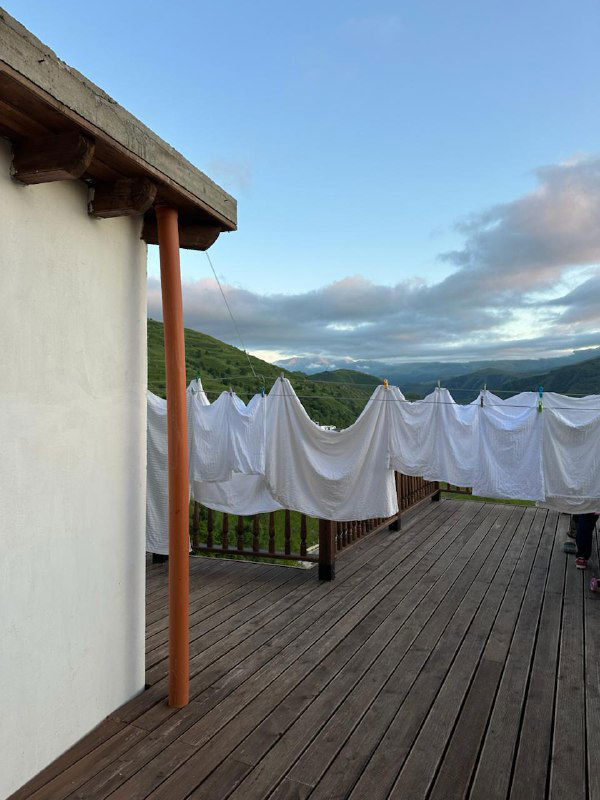 Деревянная терраса кафе с белыми простынями на верёвке, ограждением и удалённым горным хребтом под голубым небом.