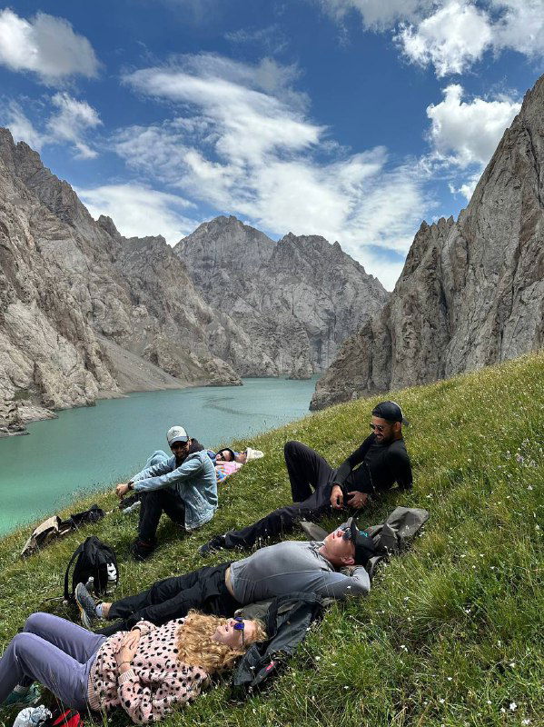 Группа туристов отдыхает на травяном склоне у бирюзового озера в ущелье Тянь‑Шаня; рюкзаки и спокойная горная панорама вокруг.