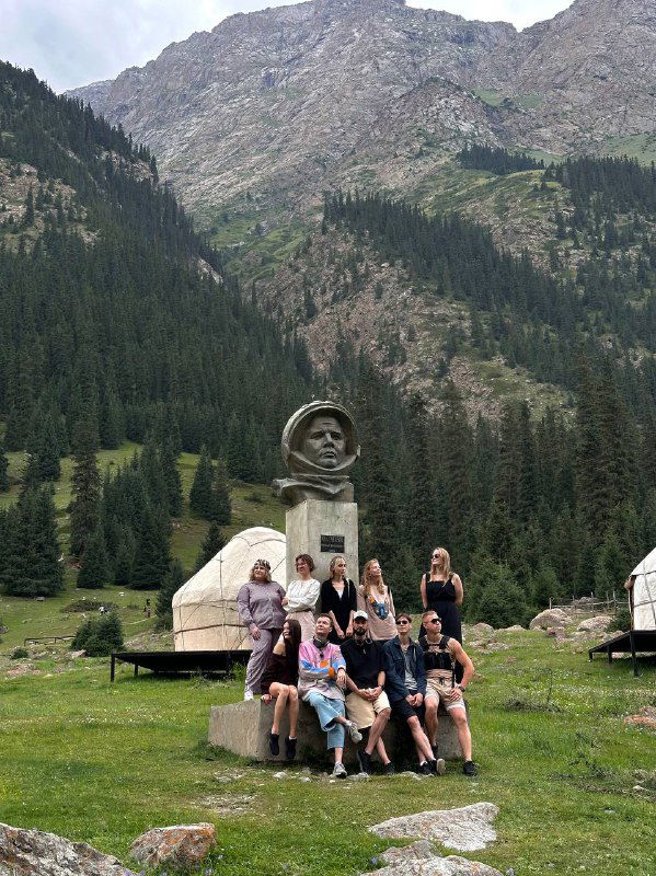 Группа путешественников у памятника на зелёной поляне рядом с юртами; на фоне — лесистые склоны и горные вершины, характерные для региона.