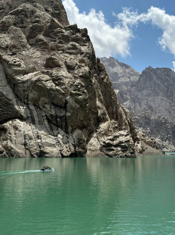 Бирюзовое горное озеро, окружённое отвесными скалами; спокойная гладь воды и маленькая лодка у подножия внушительных утёсов вдалеке.