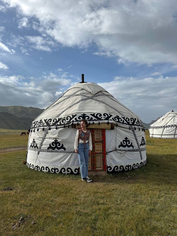 Традиционная белая юрта с декоративным орнаментом на степной поляне; турист у входа на фоне широкого горизонта и холмов Кыргызстана.