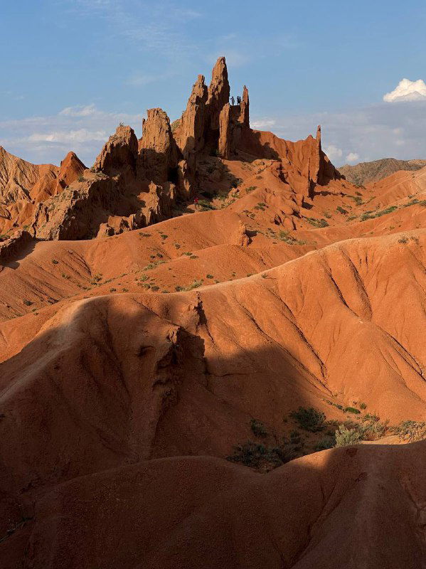 Высокие оранжево‑красные скалистые образования и колонны в полупустынном горном ландшафте Кыргызстана; выразительная геология и текстура пород.