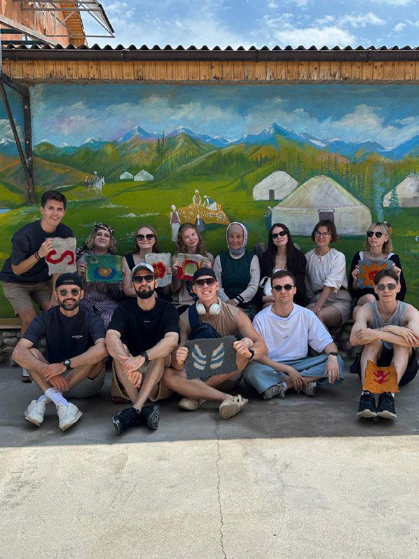Группа путешественников на фоне раскрашенной стены с изображением гор и юрт — совместное фото на фоне местного арт-оформления в селе.