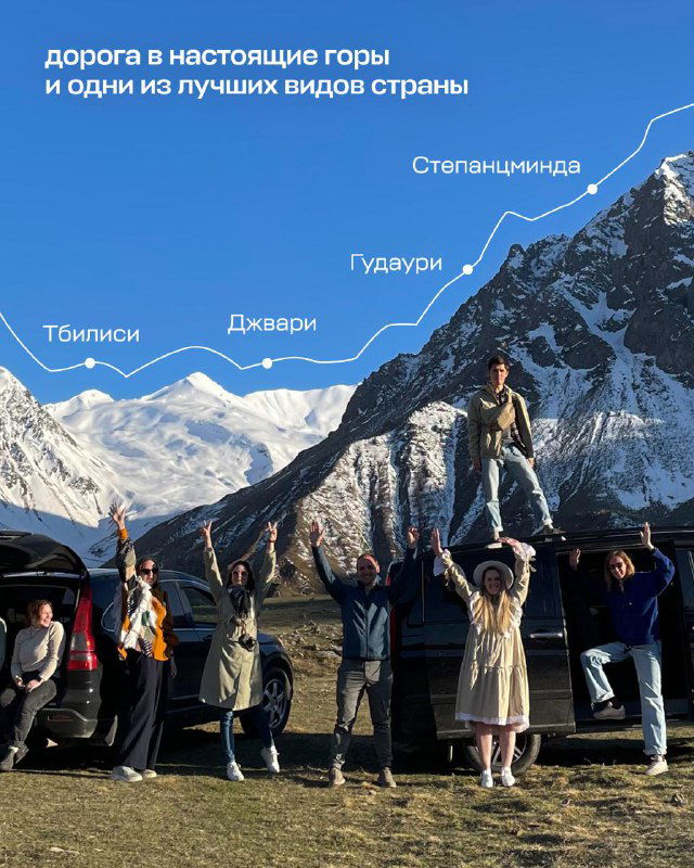 Группа путешественников на фоне заснеженных вершин в Степанцминде; люди указывают на горы, походная атмосфера и вид на Казбек.