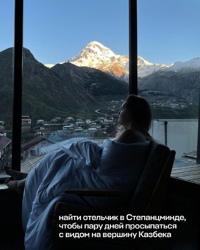 Утренний вид из окна номера с силуэтом туриста на кресле и панорамой горного поселка у подножия заснеженной вершины.