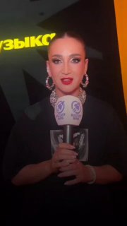 Крупный план ведущей в платье с ярким макияжем и серьгами: она держит микрофон с логотипом на фоне сцены и баннера.