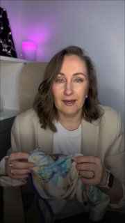 Женщина средних лет в офисе говорит в камеру, держит платок или ткань в руках; мягкое освещение, розовая лампа и полка с декором на заднем плане.