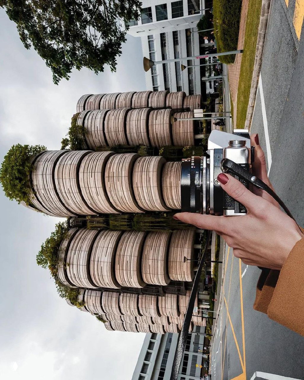 Рука держит винтажный фотоаппарат на переднем плане; на заднем плане цилиндрические деревянные башни и улица — перспективная композиция.