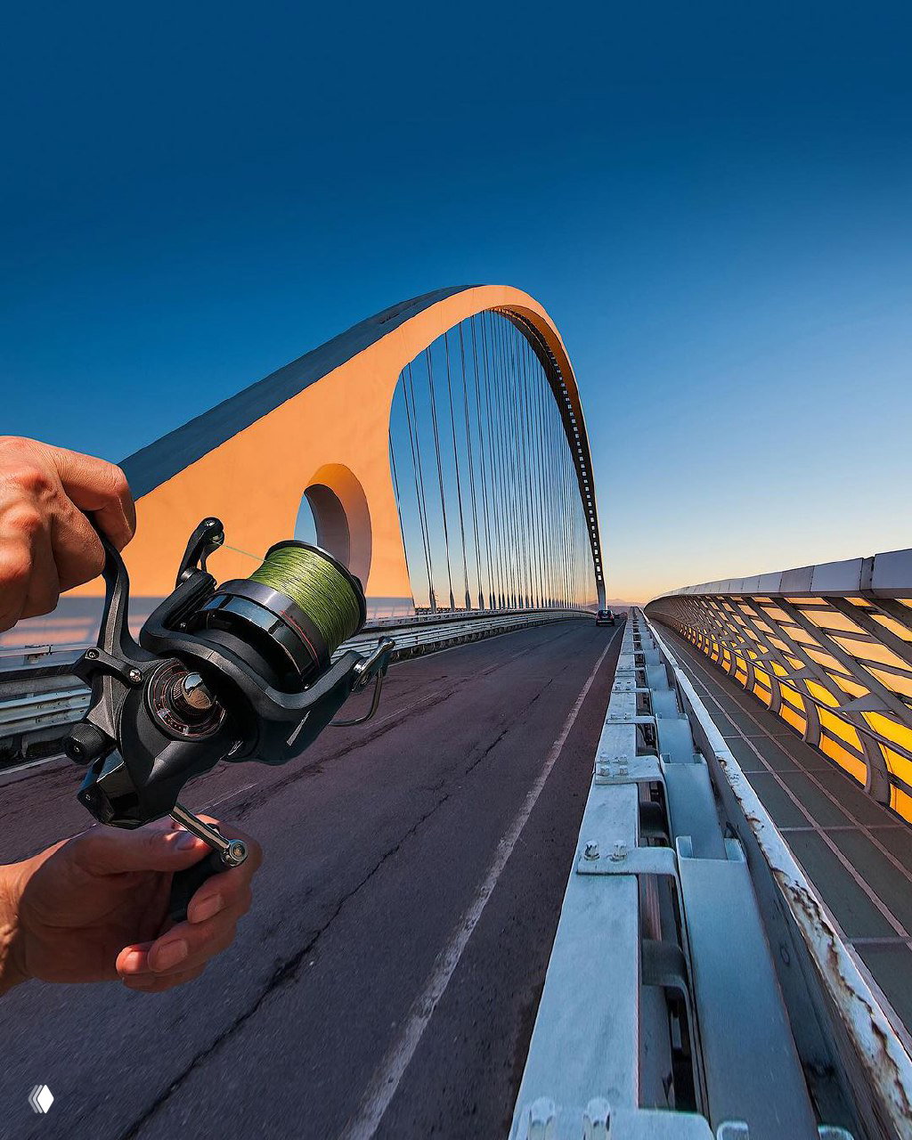 Рука с катушкой для рыбалки на фоне современного арочного моста на рассвете или закате — сочетание хобби и городской архитектуры в одном кадре.