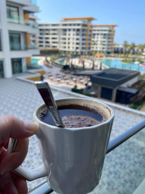 Рука держит чашку кофе на фоне комплекса отеля с бассейном и шезлонгами — утренний кадр отдыха и рабочей паузы в отпуске.