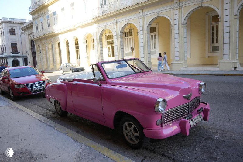 Розовый классический автомобиль припаркован у желтого здания в Гаване — вид на переднюю решётку, фары и городской фон.