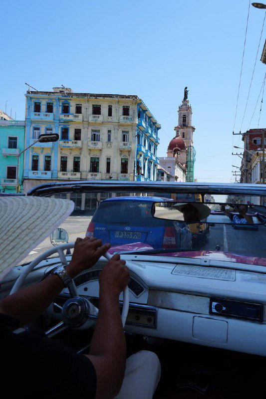 Вид из салона старого автомобиля: руки за рулём, открытая крыша кабриолета, улицы Гаваны и разноцветные фасады домов.