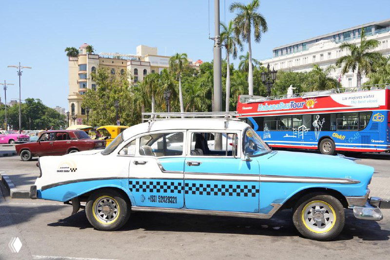 Голубо-белая ретро-машина такси в Гаване среди других старинных автомобилей — фасад здания и уличная сцена создают атмосферу города.